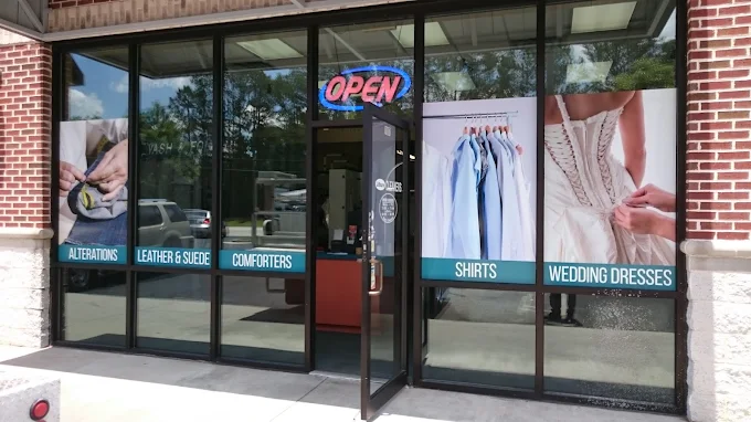 Storefront with large windows displaying images and text for alterations, leather & suede, comforters, shirts, and wedding dresses; door is open and a lit 