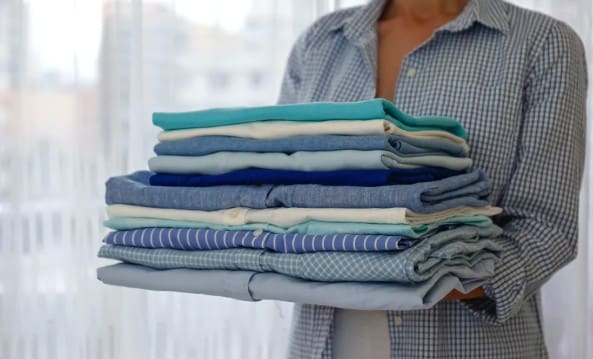 A person in a checkered shirt holds a neatly folded stack of assorted blue and white clothes against a light background.